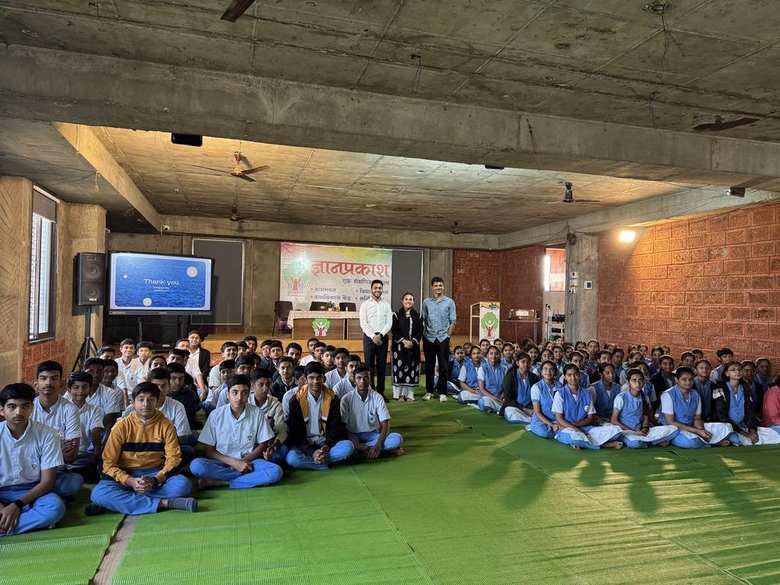 Full group of students seated on the floor with Neha and Hriday standing at the front of the hall